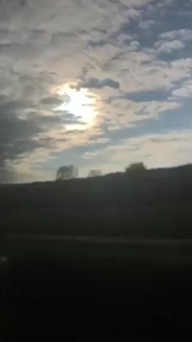 A camera records a blurry scene from the inside of a moving vehicle. The sun is shining brightly through patchy clouds in a blue sky, creating a halo effect. A dark, silhouetted hill line with a few trees is visible in the distance. The road is black. Part of the vehicle's interior is seen at the top of the frame.