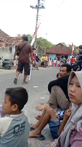 The video shows a street scene with people sitting on the ground, watching an event. In the foreground, a young child wearing a grey shirt with "HOCKEY" written on it is visible. Next to the child is a woman wearing a hijab. To their right, a man in a black shirt and beige pants is seated, looking towards the street. A young girl in a green shirt and black hijab is also present. Further down the street, several people are gathered, and a man wearing a brown shirt and shorts is walking away from the camera, holding a white pole. Indonesian flags are visible in the background, along with houses and parked motorcycles. The overall impression is of a local gathering or celebration.