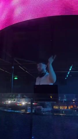 A man in a white t-shirt, DJing, stands behind a DJ deck in a club. He’s wearing headphones around his neck. A laptop sits on the deck. He smiles and raises his arm with his index finger pointed upward. The club is dimly lit with colored lights in the ceiling and background. There are several rows of lights behind the deck.