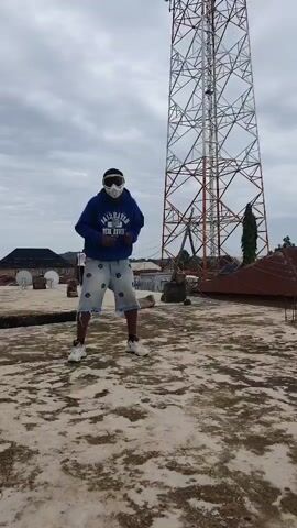 The media shows a person wearing a blue hoodie, light-colored shorts with a pattern, white sneakers, and goggles or a face mask. They are standing on a flat, outdoor surface, possibly a rooftop, with a grey, cloudy sky above. In the background, a large, metal lattice tower, likely a telecommunications tower, dominates the scene. To the left, a building with a dark roof and some satellite dishes is visible. Further in the distance, there are more buildings and some trees. The ground surface is a textured concrete or asphalt.

The person appears to be engaged in some form of dance or movement, with their arms bent and body positioned as if mid-motion. The overall mood is somewhat obscured by the mask/goggles, but the pose suggests active engagement. The weather appears overcast, suggesting it could be daytime, though the specific time is not discernible. The presence of the telecommunications tower and the style of buildings hint at a semi-urban or rural environment.

Visible text on the hoodie reads "FAYETTEVILLE" above "BLUE DEVILS".
