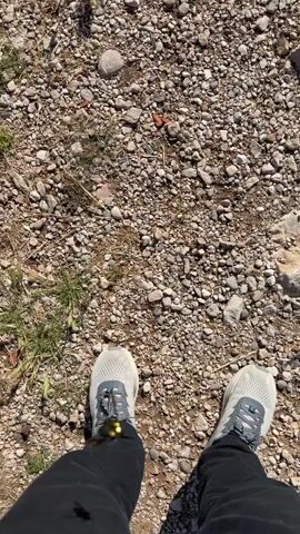 This is a top-down video clip of a person's feet standing on a gravelly path. The person is wearing black pants and light grey athletic shoes with blue accents. The ground is covered in small, greyish-brown pebbles, with sparse patches of dry grass and some larger rocks scattered around. The scene appears to be outdoors in daylight, likely a rural or natural setting given the unpaved path and natural ground cover. There are no discernible emotions or specific events taking place beyond standing still. The time of day appears to be daytime, possibly late morning or afternoon, based on the shadows cast. The location is identified as Ponikvica, Montenegro, which aligns with the natural, somewhat rugged terrain depicted. There is no visible text in the video.