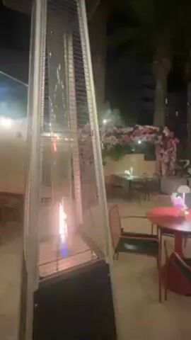 The video shows an outdoor restaurant patio at night. A tall, pyramid-shaped patio heater with a visible flame is on the left side of the frame. Moving toward the right, there is a round red table with three chairs around it. In the background, there is a decorated archway of pink flowers. There are other tables and chairs further back on the patio. Palm trees are visible in the background. Cars drive by in the distance.