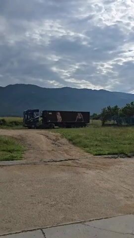 The camera zooms in on a large cargo truck with a dark brown trailer parked in a field with green grass and a dirt road. Two large images of women are displayed on the trailer. The woman on the left is wearing a nude-colored bra, while the woman on the right is wearing a black bra and camisole. In the background, there are mountains and a cloudy sky.