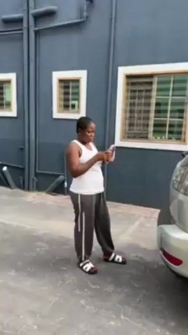 A dark-skinned woman with braided hair stands outside near a dark blue building with three windows. She is wearing a white tank top, dark gray sweatpants, and sandals. She looks intently at a cell phone in her hands. She has a string of pink beads looped around her wrist. A gray vehicle is parked to her right. The building has visible pipes and electrical conduits.