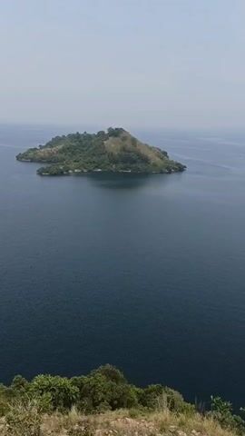 The video opens with a wide shot of a verdant, hilly island surrounded by deep blue water. The camera slowly pans right, revealing two more islands in the distance, partially obscured by a hazy sky. The scene is shot from a high vantage point, with foliage visible in the foreground, suggesting the camera is positioned on a hill or cliff. The water appears calm with small ripples on its surface. The overall impression is one of a serene, natural landscape.