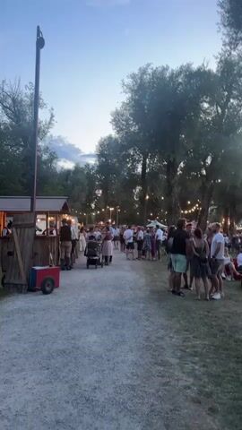 The video shows an outdoor gathering, possibly a festival or market, near a body of water. In the foreground, a gravel path is lined with people, some standing in groups and others walking. To the left, a wooden stall with a red and blue cart is set up, and a person in traditional German attire is visible behind the counter. String lights are strung between trees in the background, illuminating the scene as dusk settles. Further back, several boats are docked on the water, reflecting the soft glow of the lights. The overall atmosphere appears relaxed and festive.