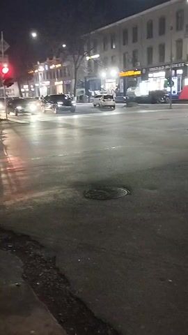 The video shows a nighttime intersection scene. Cars move through the wet streets with their headlights reflecting on the pavement. Buildings line the streets with lighted storefronts. The traffic light is red in one direction. A manhole cover sits in the road.