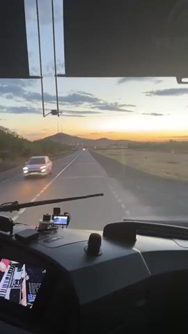 This video shows the view from the cockpit of a vehicle, likely a bus or truck, driving on a highway towards a sunset. The sun is setting behind distant hills, casting a warm orange and yellow glow across the sky, which is filled with scattered clouds. The road stretches out ahead, with white lane markings indicating it's a highway. Trees line the left side of the road, and flat, grassy fields stretch out to the right.

Inside the vehicle, we can see part of the dashboard. Mounted on the dashboard are various devices, including what appears to be a GPS or navigation screen and possibly a dashcam. To the left, a screen displays a visualizer or graphic related to music, showing what looks like a keyboard interface with musical notes.

In the foreground, a white car is passing the vehicle, its motion creating a blur. The time of day is clearly sunset, as indicated by the warm lighting and the position of the sun near the horizon. The weather appears clear with some clouds. The setting is a rural highway in Kazakhstan, given the landscape and the visible text "YUTO" on the interior of the vehicle, which is a known bus manufacturer in the region. The overall mood is one of travel and perhaps peaceful contemplation as the day ends.