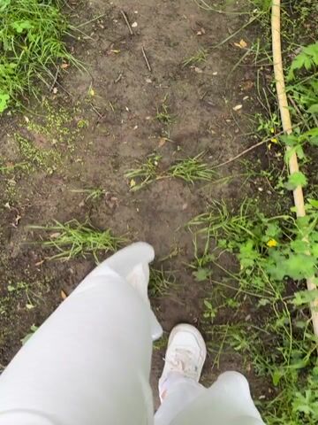 The video provides a first-person perspective of someone walking along a narrow, damp dirt path. The person is wearing white sneakers and white, loose-fitting pants. The path is surrounded by vibrant green grass and various small plants. A long, light-colored wooden stick or branch lies parallel to the path on the right side.

Around the 0:04 mark, the camera pans upwards, revealing more of the environment. The dirt path continues through an area densely covered with lush green foliage. In the background, a wooden structure, possibly a house or cabin, is visible with a porch and wooden stairs. Several thin tree trunks stand between the path and the building. A small metal chair or table can be partially seen on the grass near the structure. The setting appears to be a natural, semi-developed outdoor space, possibly a garden or a wooded area adjacent to a residence, consistent with a rural or suburban location like Vozhoiskii, Russia. The lighting suggests an overcast day, creating a soft, natural ambiance. The primary activity is walking and observing the natural surroundings. No visible text is present.