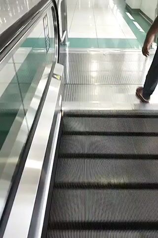 The video shows an indoor moving walkway with people walking on it. The walkway is in a modern building with bright lighting. There is signage in Arabic and English, indicating "Red Line". The camera appears to be moving, possibly being carried by someone walking on the walkway. The scene is clean and well-maintained.