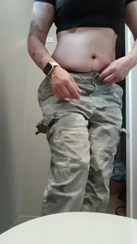 A woman wearing a black crop top and camouflage cargo pants is standing in a bathroom, facing the camera. She fastens the button on her pants. She turns to her left and rests her left hand on the pants.  A white bathroom sink is in the foreground. A doorway is visible behind her.  The lighting is bright.  She has tattoos on her left arm and chest.
