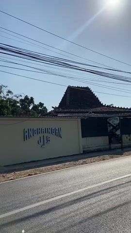 The video begins with a view of a building with the word ANGKRIINGAN written on it in stylized white letters. The building has a traditional Indonesian tiled roof. A road is visible in the foreground, and power lines stretch across the sky. The sun is shining brightly, casting shadows. The camera then pans to the right, showing a wall with a sign that reads "BUKA JAM 09.00 WIB". Further down the road, a person is riding a bicycle, followed by several motorcycles. The scene appears to be a roadside in a rural or semi-rural area, with trees and vegetation on either side of the road. The sky is clear and blue.