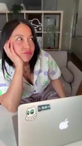 A woman sits at a table with a laptop in front of her. She rests her head on her hand and looks bored, but when someone dangles a green stretchy toy in front of her, she smiles and grabs at it. She then puts the toy on her face while music plays in the background. The woman is wearing a short-sleeved collared shirt with marijuana leaf patterns and sweatpants. The laptop is grey with an Apple logo and stickers. The room has a grey chair, white shelves, and a glimpse of a kitchen.