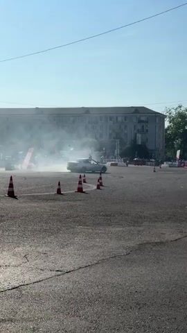 The video shows a white car drifting in a parking lot or an empty street. There are orange cones set up in a pattern on the ground, indicating a course for the drifting event. Smoke is coming from the tires of the car as it slides around the track. In the background, there is a large building and some trees. There are also spectators visible in the distance, as well as some banners and tents. The sky is clear and blue, and the sun is shining. The overall impression is of a car drifting event or competition taking place outdoors on a sunny day.