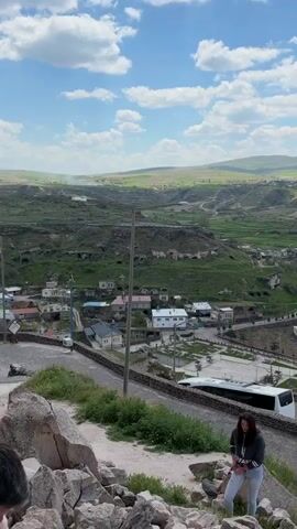 The video captures a panoramic view of a village and its surrounding landscape in Acıgöl, Turkey, under a bright, partly cloudy to clear blue sky.

The initial shot frames a village nestled in a valley, featuring houses with light-colored roofs. The hillsides above the village display numerous ancient cave dwellings carved into the rock, interspersed with green grass. A street light pole is visible in the foreground.

As the camera pans right, it reveals extensive green, terraced agricultural fields and a winding road that stretches across the landscape. A red flag, likely the Turkish flag, is visible on a distant hill. The sweep continues to showcase a vast, open plain with more fields and distant settlements.

The final moments of the video focus on the rocky cliff face, highlighting several prominent cave entrances and openings, suggesting a history of human activity within these natural formations. Green vegetation and scattered rocks are visible at the base of the cliffs. No people or discernible text are present in the video.