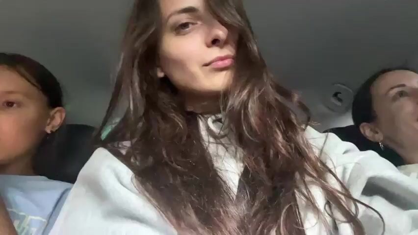 The video shows a close-up of a young woman with long brown hair in the front passenger seat of a car. She is looking at the camera and making a kissing face. Her eyes are blue, and she is wearing a gray hoodie. In the background, another young woman is visible in the driver's seat, and a child is in the back seat. The car appears to be filled with luggage. The woman in the foreground speaks Russian, stating "No, no, as I say, all in all without adventures."
