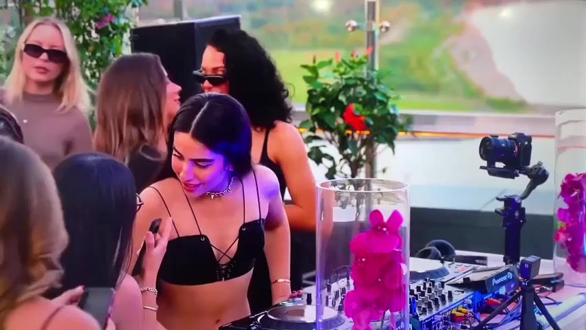 A group of women are gathered around a DJ setup at an outdoor event. The DJ, a young woman with long dark hair and a black top, is smiling and interacting with a woman holding a phone. Behind her, a woman in sunglasses watches. The DJ setup is decorated with flowers in tall glass cylinders, and a camera is mounted on a tripod nearby. In the background, a cityscape can be seen.