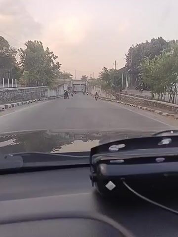 The video shows a view from inside a car driving on a road. The road is paved and has concrete barriers on both sides. There are several motorcycles on the road, some in front of the car and some in the distance. Trees and buildings are visible along the sides of the road. The sky is overcast. The time stamp in the video indicates it was recorded at 00:00:00 to 00:00:04. A black device with lights is visible on the dashboard of the car. The car appears to be approaching an overpass.