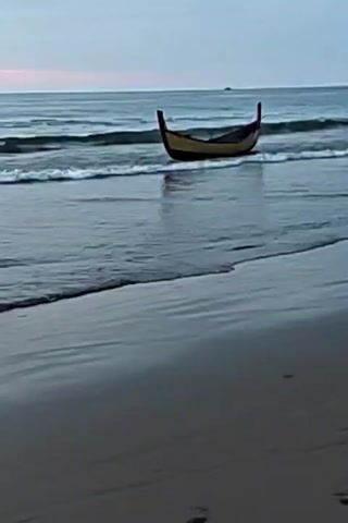 A long, narrow boat with a curved bow and stern sits in shallow water near a sandy beach. The boat is light-colored with a dark stripe along its side. Waves gently lap around the boat and wash onto the shore. The sky above is overcast with a hint of pink on the horizon, suggesting dawn or dusk. The overall mood is tranquil.