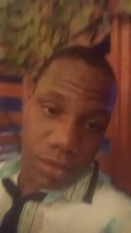 A close-up video shows a man with dark skin wearing a light-colored collared shirt with a green and white pattern. He has a dark tie. He is smiling and looking at the camera. The background is blurry and appears to be outdoors with some foliage and a wooden structure. There is a fan visible in the background to the left.