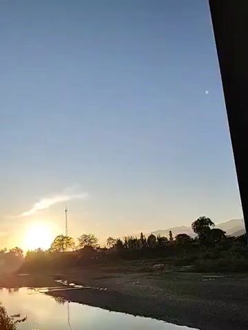 The video shows a serene landscape at sunset or sunrise. A calm river flows in the foreground, reflecting the golden light of the setting sun. The horizon is marked by trees and the silhouette of a mosque's dome. A tall communications tower stands among the trees. The sky is a gradient of blue, transitioning to a warmer hue near the sun. A single, bright celestial object, possibly the moon or a planet, is visible in the upper right of the sky. The camera is static, offering a steady view of the scene.