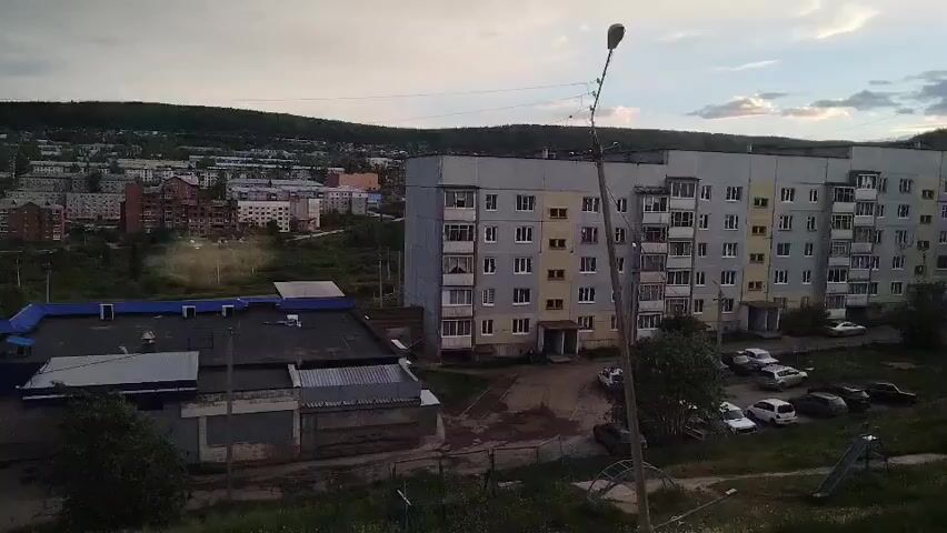 This media file presents an outdoor scene in Zheleznogorsk-Ilimsky, Russia. The primary subjects are residential buildings and a low-rise commercial structure, interspersed with parked cars and vegetation. The scene is dominated by a multi-story apartment building, characterized by its gray facade and numerous windows. Adjacent to it is a single-story building with a blue roof, likely a shop or public facility. Several cars are visible in the parking area between the buildings, and a children's slide is situated on the right side of the frame.

The setting appears to be a residential neighborhood. The background shows a more densely populated area with various other buildings, suggesting a town or city environment. Rolling hills covered in vegetation form the horizon, indicating a natural landscape surrounding the settlement.

The time of day is likely late afternoon or early evening, as indicated by the soft, diffused light and the presence of clouds in the sky, suggesting overcast or partly cloudy weather. The overall atmosphere is calm and ordinary, capturing a typical day in this Russian locale. There is no visible text within the media file.