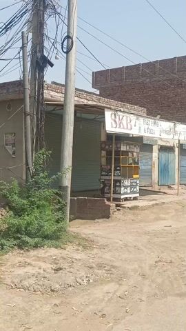 The video shows a storefront called "SKB Fast Food & Pizza" located on a dirt road in what appears to be a rural area. The store has a painted sign above it and what looks like a mobile food stand in front, also labeled "SKB Foods & Pizza." The storefront has a closed metal shutter. A person is seen standing near the entrance of the store in the background. The weather looks clear and sunny. The buildings surrounding the storefront appear to be constructed of brick.
