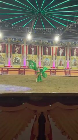 A young woman in a shimmering green costume, possibly a dancer or performer, is shown in the center of a sandy arena. She is spinning and twirling, her arms outstretched, causing large, flowing green fabric attached to her costume to billow around her. The fabric appears to be two large pieces, one extending behind her and the other to her side, creating a dramatic effect. She has her hair pulled back. The background suggests a theatrical or circus-like setting, with a large, ornate backdrop depicting classical Roman-style architecture with statues and painted murals. The murals appear to be of mythological or historical scenes. There are columns and a red and yellow striped border along the base of the backdrop. Colored lights, primarily pink, illuminate parts of the background. The floor of the arena is covered in sand, and there are spotlights casting bright yellow light on the sand near the performer. In the foreground, a dark, blurry area suggests the edge of seating or a stage.