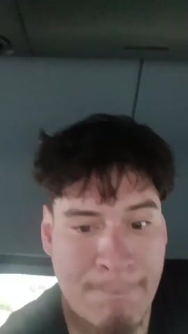 A head-and-shoulders shot shows a man in what appears to be a vehicle with a dark headliner. The man has short, dark hair with bangs. He looks down at the camera, his mouth moving as if speaking. The background is blurry.