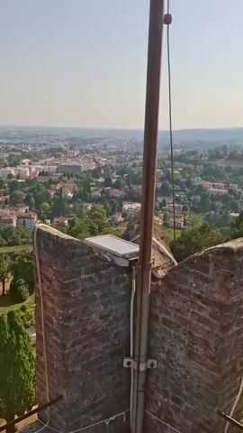 A camera pans from a brick structure with a light fixture attached, revealing a panoramic view of a European city with red-tiled roofs and green trees under a clear blue sky. The camera then moves to show an Italian flag flying from a flagpole, with a telescope positioned in the foreground. The video captures the cityscape, lush greenery, and a national symbol, suggesting a scenic overlook or historical site.