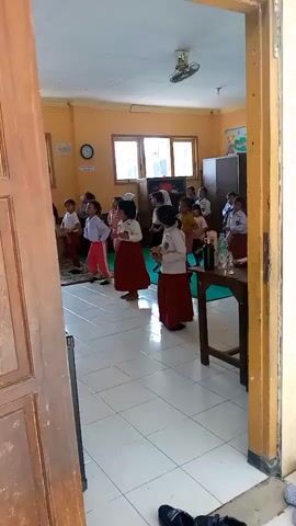 The video shows a group of young children, likely students, in what appears to be a classroom. They are dressed in school uniforms, consisting of white shirts and maroon skirts or pants. Most of the children are standing in rows, facing the front of the room, and appear to be engaged in some sort of activity, possibly singing or a physical exercise, as some are moving their arms. There is a teacher or adult figure standing at the front, on the right side of the frame, also participating. The classroom has yellow walls, windows with blinds, a ceiling fan, a clock on the wall, and a blackboard at the back. There are also desks and chairs visible, as well as a small table with bottles of water and a book. The floor is tiled. The scene is captured from the doorway of the room.