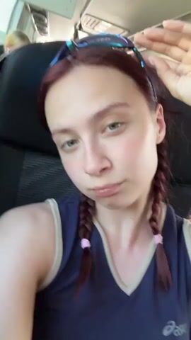 A woman with red hair and pigtails, wearing a navy blue top, is sitting on a bus. She looks at the camera with a neutral expression and then puts on a pair of blue reflective sunglasses. The inside of the bus is visible in the reflection on the sunglasses.