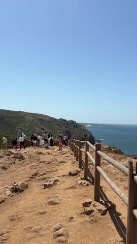 This media file captures a scenic view from a cliffside overlooking the ocean. In the foreground, a rustic wooden fence with two horizontal rails runs along the edge of the cliff. The ground is a mixture of dry, reddish-brown earth and scattered rocks.

Further back, a group of people are gathered along the cliff path, some standing and looking out at the sea, others walking. They appear to be tourists enjoying the vista. The setting is a coastal landscape with rolling green hills on the left side of the frame, meeting the steep cliff face.

The ocean is a deep blue, stretching out to the horizon under a clear, bright sky. The sun is high, suggesting it is midday. The weather appears to be warm and sunny, with no clouds visible.

The location is identified as Colares, Portugal, which is known for its dramatic coastal cliffs and views of the Atlantic Ocean. The overall emotion conveyed is one of peaceful enjoyment of nature and a sense of awe inspired by the expansive ocean view. There is no visible text in the media file.