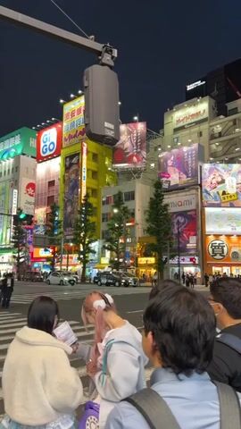 The video shows a street scene at night in Akihabara, Tokyo, Japan. Numerous bright, illuminated billboards with anime and manga advertisements tower over the street, creating a vibrant and busy atmosphere. Several people are walking across a crosswalk in the foreground, some with backpacks, indicating they are likely tourists or shoppers. The street appears to be moderately busy with pedestrian and vehicular traffic. The sky is dark, suggesting it is nighttime. The overall impression is of a lively entertainment district.