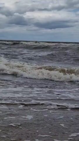 The video shows a close-up view of ocean waves crashing on a sandy beach under a cloudy sky. The water is dark and choppy, with white foam from the breaking waves. The sky is filled with grey clouds, with glimpses of blue sky peeking through. The scene appears to be during the day. There are no people or man-made objects visible in the video.