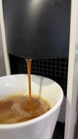 A close-up shot shows a black espresso machine dispensing a stream of coffee into a white mug. The mug already contains some coffee and foam. The background of the espresso machine is white.