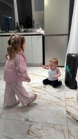 The video shows a toddler, dressed in a white tank top and dark pants, crawling on a marble-patterned floor towards a large black speaker with colorful lights. Standing near the toddler is a young girl with pigtails, wearing a pink and white striped pajama set, smiling and laughing. The scene takes place in a kitchen, with white cabinets and stainless steel appliances visible in the background. The audio suggests that music is playing. The children seem to be enjoying themselves in a domestic environment.