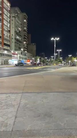 The video presents a nighttime urban street scene, likely in Puerto La Cruz, Venezuela, as the camera slowly pans from left to right. The foreground features a wide, paved pedestrian area or sidewalk.

A multi-lane road dominates the middle ground, where numerous vehicles are in motion, their headlights and taillights appearing as blurred streaks of white and red against the dark asphalt. Tall, multi-story buildings line both sides of the street. On the left, one building prominently displays a large, vibrant, and abstract mural or advertisement that glows with various colors. Other buildings have illuminated windows, indicating ongoing activity.

The scene is well-lit by streetlights, with several bright LED fixtures visible along the road and in the pedestrian areas. One distinct white streetlight with a curved arm and a bright bulb is prominent in the foreground on the right side. In the background, beyond the immediate street, more buildings and some light-colored trees are faintly discernible. A low barrier or planter separates the road from the pedestrian zone, and a white bench or seating area is partially visible on the far right. The sky is dark, confirming the nighttime setting. No discernible text is visible on any signs or buildings.