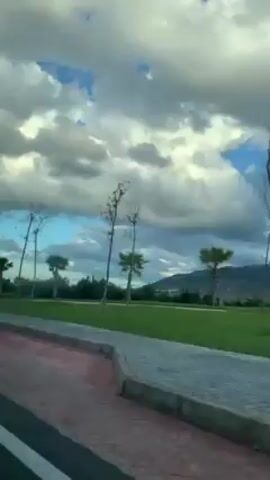 The camera appears to be moving inside a vehicle, capturing the outside view. A red-colored pavement runs alongside a grassy area with a few trees, including some palm trees. In the background, there are hills. The sky above is mostly cloudy with some patches of blue. Some structures can be seen intermittently as the camera moves, including a small white building and what looks like a lamp post.