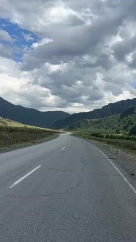 The video shows a straight, paved road cutting through a valley between green, forested mountains. The sky is filled with dramatic, dark gray clouds interspersed with patches of blue. The road has some cracks and a white line painted down the middle. The surrounding landscape includes grassy areas near the road and trees climbing up the slopes of the mountains. The overall impression is of a scenic, somewhat remote location.