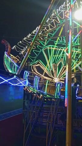 Here is a description of the video:

The video shows a carnival ride illuminated with colorful LED lights at night. The ride is constructed of metal scaffolding with designs outlined in various colors. There is a staircase leading up to the base of the ride. The light is moving and flashing and there is electronic music playing in the background. The video is shot from a low angle, looking up at the ride.