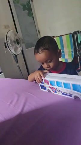 A young boy lies on a purple surface, looking at a toy bus. The boy is wearing a dark blue shirt with a red collar. The toy bus is white with blue and red markings. In the background, there is a fan on the left, and a rack with clothes on the right.