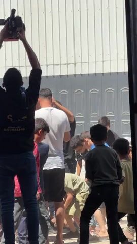 A videographer holds a camera high above their head, filming a crowd of people. Several children are visible, some wearing brightly colored shirts. One person in a pink shirt is raised on the shoulders of another person. The crowd appears to be gathered outside a building with a ribbed metal facade.