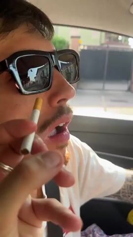 The video begins with a close-up of a person's face, holding a cigarette near their mouth. The person is wearing sunglasses and a white t-shirt. The background is slightly blurred, showing a residential area. The camera then pans to the car's dashboard, displaying a screen with an image. Following this, the perspective shifts to the side-view mirror, reflecting the person's face again, this time without sunglasses and wearing a baseball cap. The person is seen winking and holding a cigarette in their mouth, with a tattoo visible on their arm and a phone in their hand. The surrounding environment appears to be outdoors during the daytime.