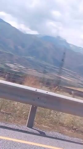 The video shows a view from a moving vehicle looking out over a landscape. In the foreground, there is a gray guardrail and a paved road with a yellow line. The landscape beyond the guardrail includes dry grass, scattered trees, hills, and mountains in the distance. There are also some man-made structures visible, including utility poles and what appear to be buildings or agricultural structures. The sky is partly cloudy. The video is shaky, indicating it was likely recorded from a moving vehicle.