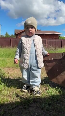 A small child is standing in a grassy yard on a sunny day. The child is wearing a light blue jumper, a beige vest, a grey beanie, and black and white shoes. The child is standing next to a rusty piece of metal, possibly part of a farm implement. In the background, there is a brown wooden fence, a house with a brown roof, and a bright blue sky with puffy white clouds. The camera zooms slightly and then focuses on blades of grass in the foreground. The child looks directly at the camera.