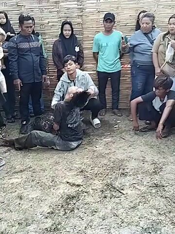 The video shows a group of people standing around two people wrestling on the ground. The wrestling is taking place on a grassy area with a woven bamboo fence in the background.  Several adults and a child are watching the wrestling match. One of the wrestlers is lying on the ground with the other on top of him, seemingly trying to subdue him. Both wrestlers are covered in dirt or mud. The people watching appear to be a mix of ages and genders. One person is kneeling near the wrestlers, possibly offering assistance or encouragement.