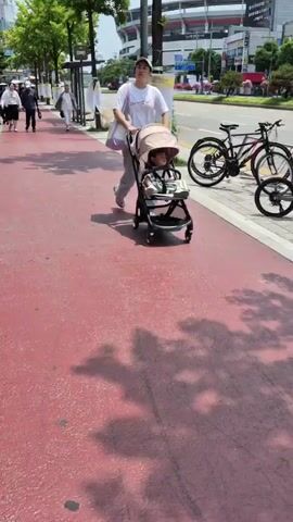 A woman pushes a stroller with a child in it on a red paved sidewalk next to a street. There are trees lining the sidewalk, and other pedestrians are walking in the background. The woman is wearing a white t-shirt and black shorts, and a black baseball cap. The child is wearing a white and blue outfit. There are bicycles parked on the other side of the sidewalk, and a stadium can be seen in the distance. The weather appears to be sunny with some clouds.