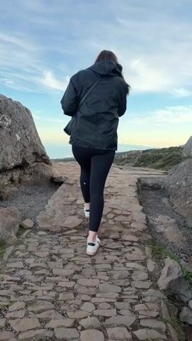 A woman walks away from the camera on a stone path. She is wearing black leggings, white sneakers, and a dark jacket. The path is made of uneven stones and surrounded by grass and rocks. Beyond the path, there is a scenic view of a mountainside covered in green vegetation, with the ocean visible in the distance under a blue sky with wispy clouds. As the woman exits the frame, the view widens to show more of the mountainside.
