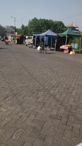 A person on a bicycle wearing a hijab and carrying a white bag is riding past a market stall. The market has blue and green tents. There are vehicles, including a van and motorcycles, parked near the stalls. The ground is paved with cobblestones. A dirt bike is visible in the foreground. The sky is clear and the sun is shining.